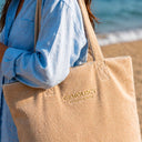 Femme portant le sac de plage beige Gemology à l'épaule au bord de la mer, logo brodé doré visible